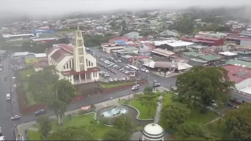 Vista aérea de un pueblo con iglesia blanca de torre alta, parque central y techos de colores bajo la neblina.